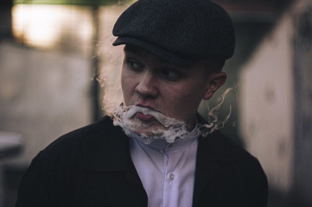 pexels-photo-10312209 A young man in a flat cap blows a smoke ring while standing outdoors. Moody and vintage style.